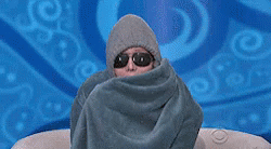 Person wrapped in a blanket with sunglasses on sits on a gray couch and pulls blanket closer