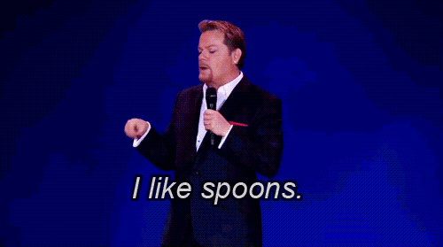 Iconic genderfluid comedian Eddie Izzard, looking dapper in a suit on a blue background, makes a scooping gesture and says "I like spoons."