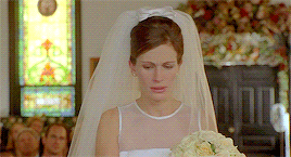 Julia Roberts, dressed as a bride at the altar, holds up a gloved hand, shakes it, and turns away