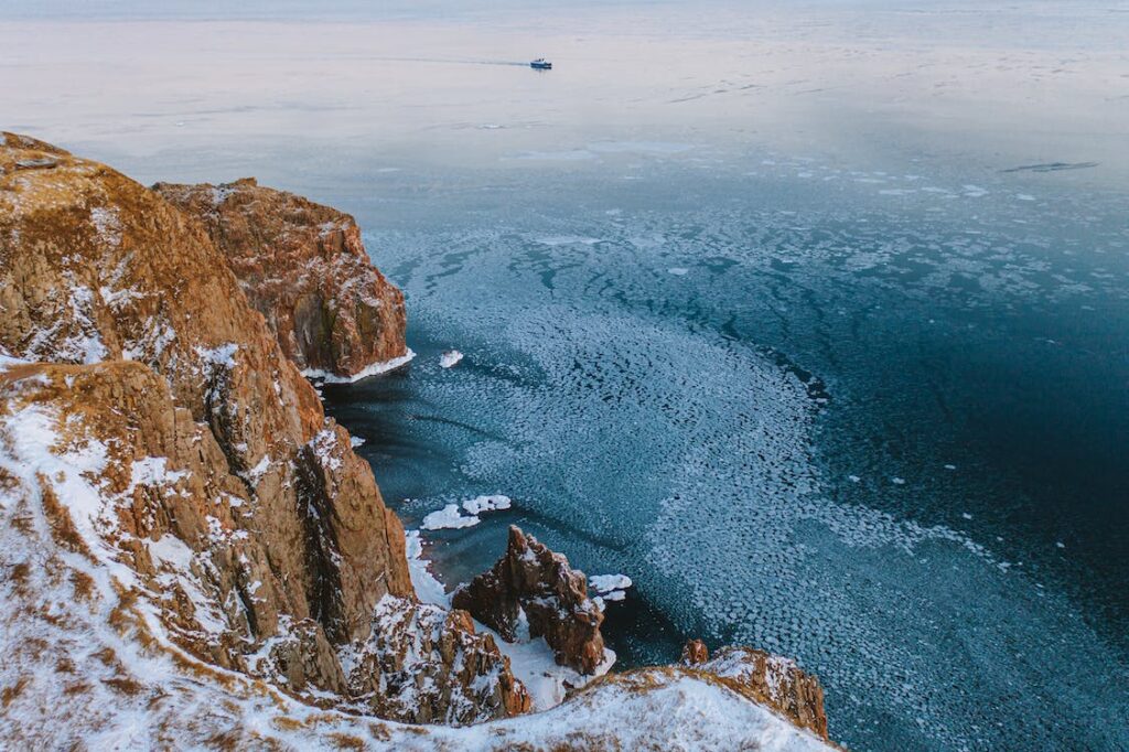 An image of bare snow-covered cliffs and icy water, with a ship approaching in the far distance.