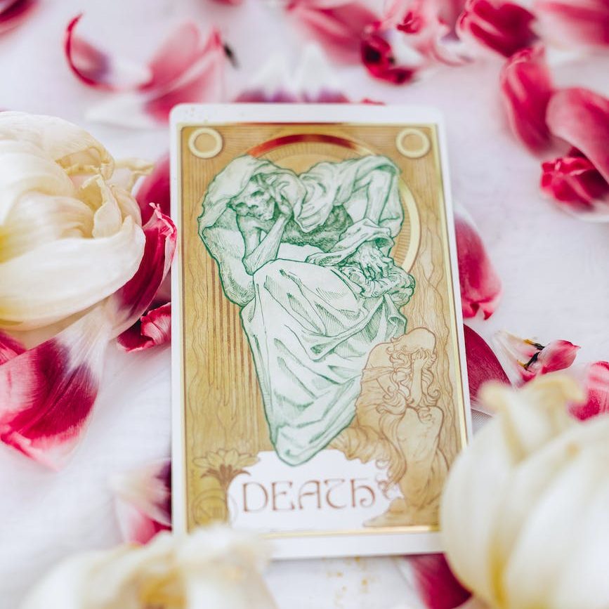 A Death tarot card with art deco style line art of the titular skeletal figure in green with ornate letters and a weeping person in gold below is displayed among pink and white petals on a white background.